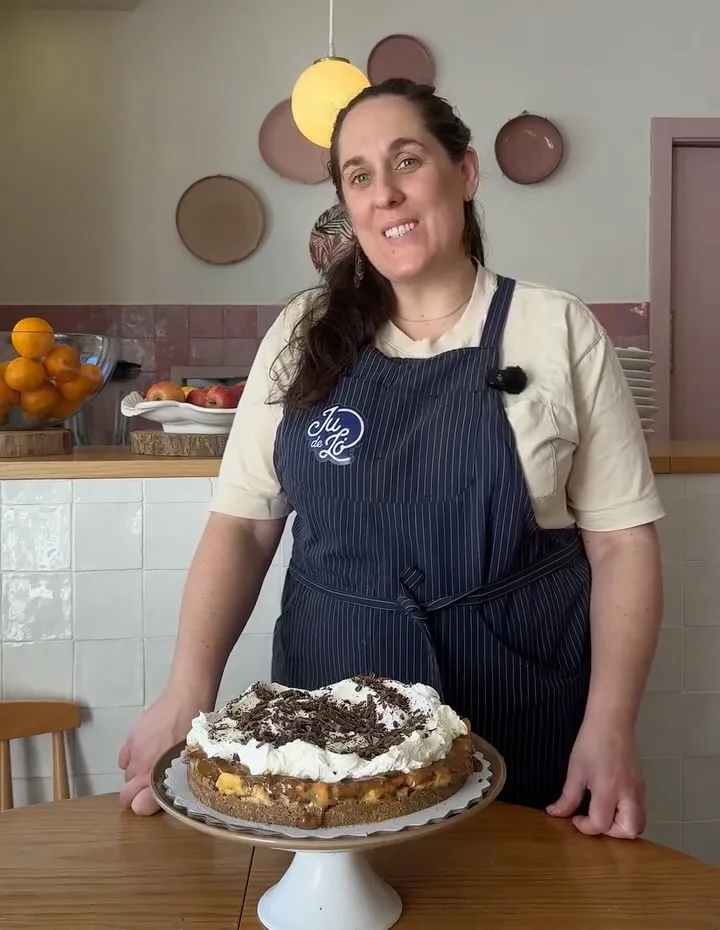 Pasteleira de avental às riscas com o logótipo Ju de Ló, a sorrir junto a uma banoffee com chantilly e raspas de chocolate, com o balcão de azulejo rosa ao fundo
