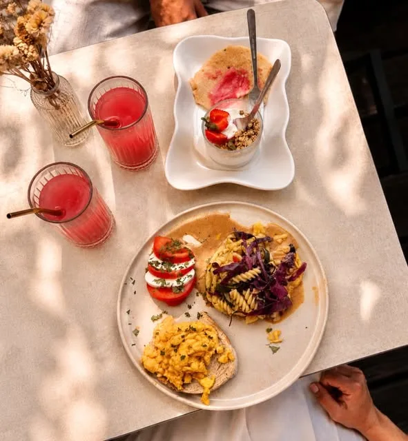 Mesa com luz filtrada, com dois sumos vermelhos, prato de brunch com ovos mexidos, tomate com mozzarella e massa, e iogurte com granola e morangos